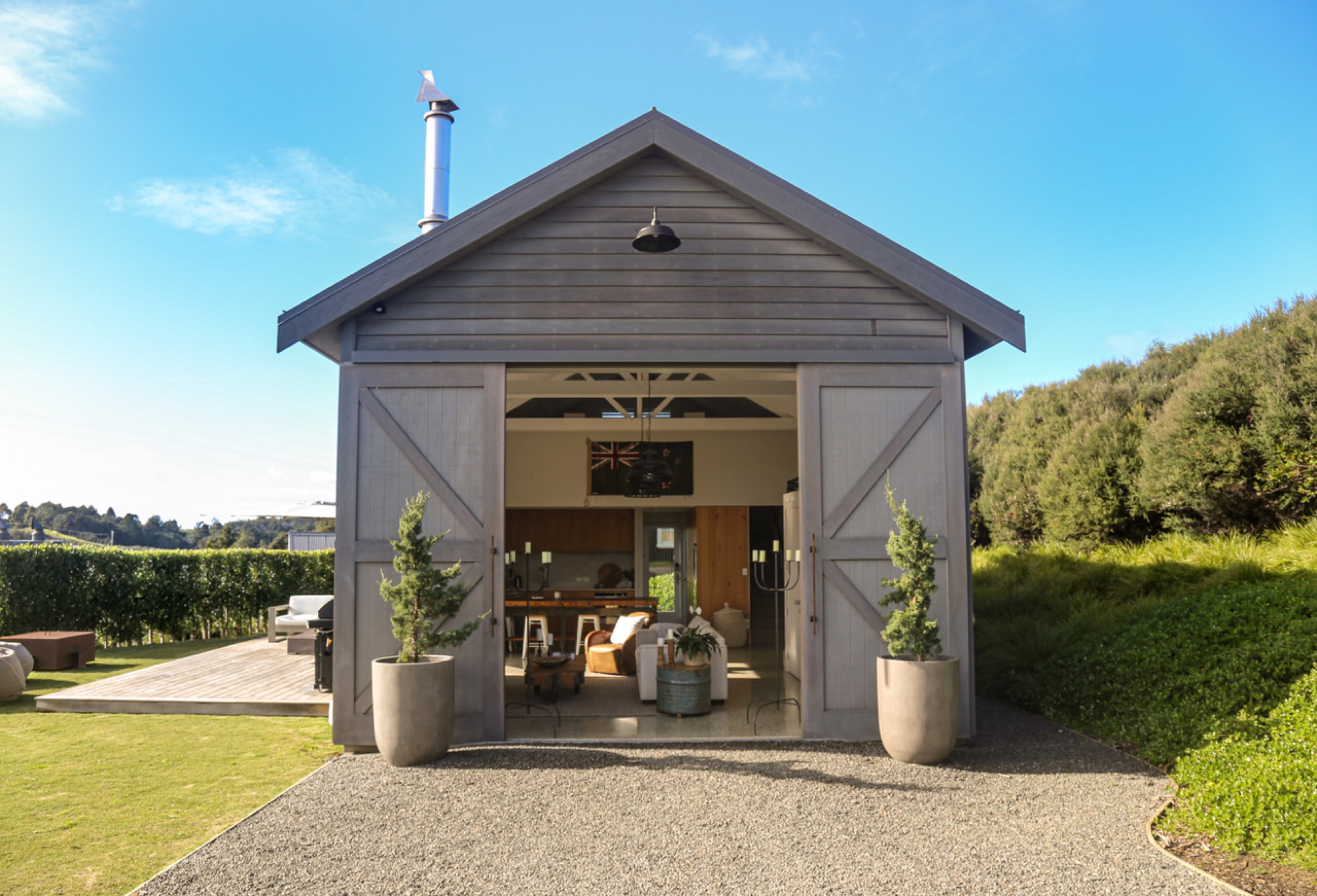 The Boat House at Bluestone Estate from the drive: grey weatherboards, the barn doors slid open to the living room, potted conifers either side, and the deck and hedge beyond.