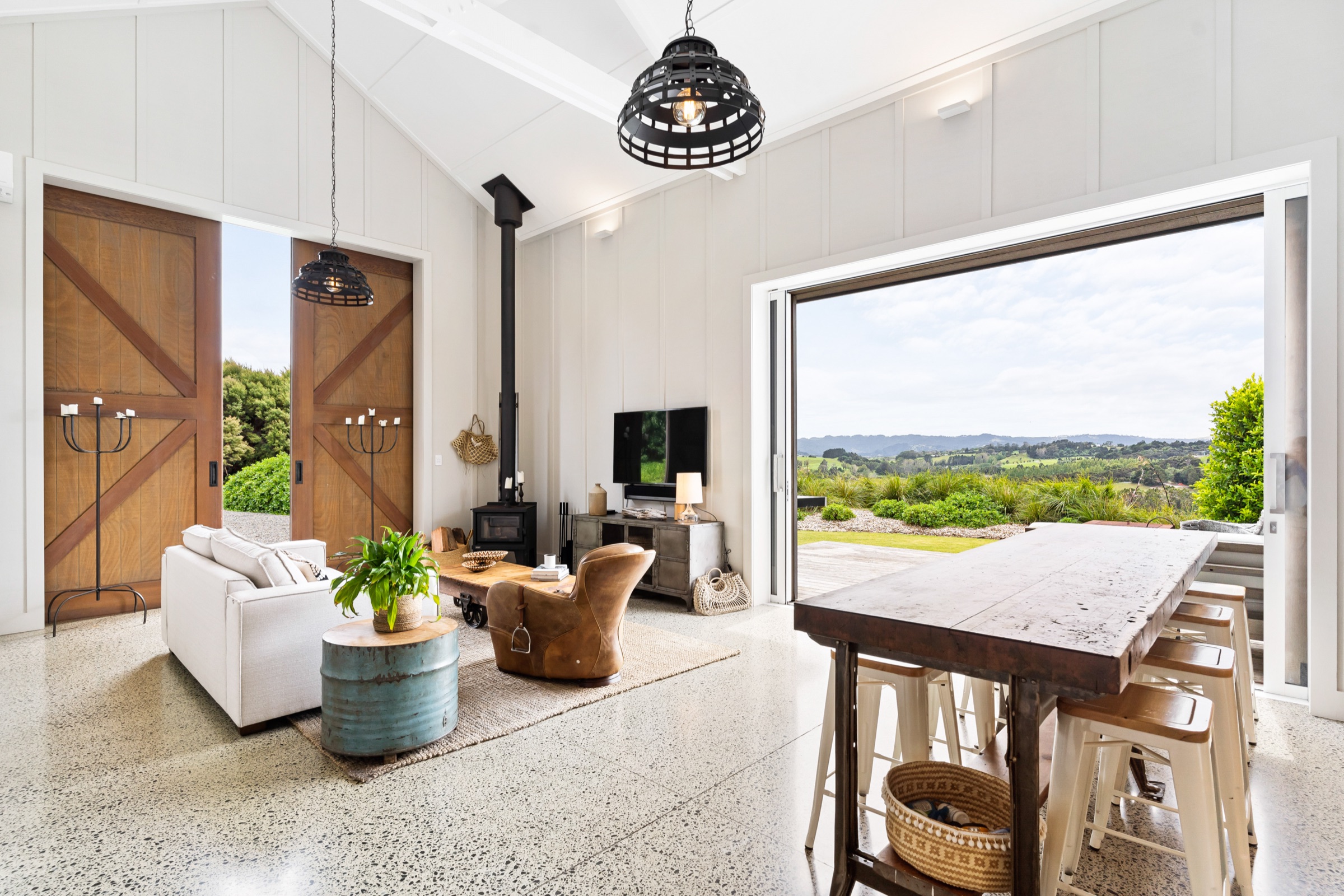 Inside the Boat House at Bluestone Estate: white-panelled walls, the pot-belly stove, a leather armchair, the long timber table with stools, and the doors open to rolling farmland.