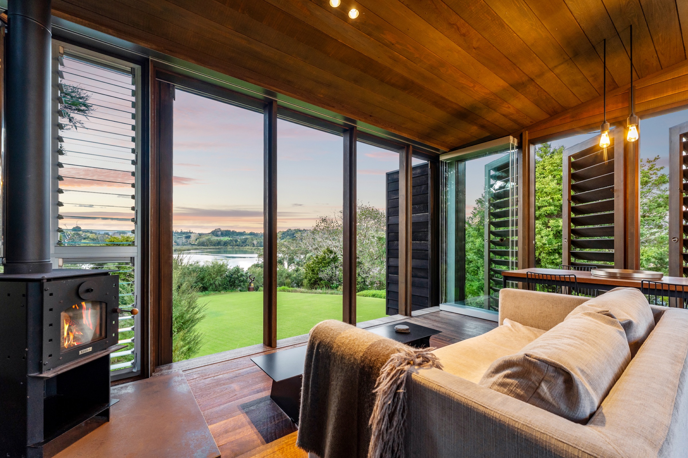 Inside the living cabin of the Cabins at Bluestone Encampment: the wood burner lit, a linen sofa, and floor-to-ceiling windows over the lawn to the estuary at sunset.