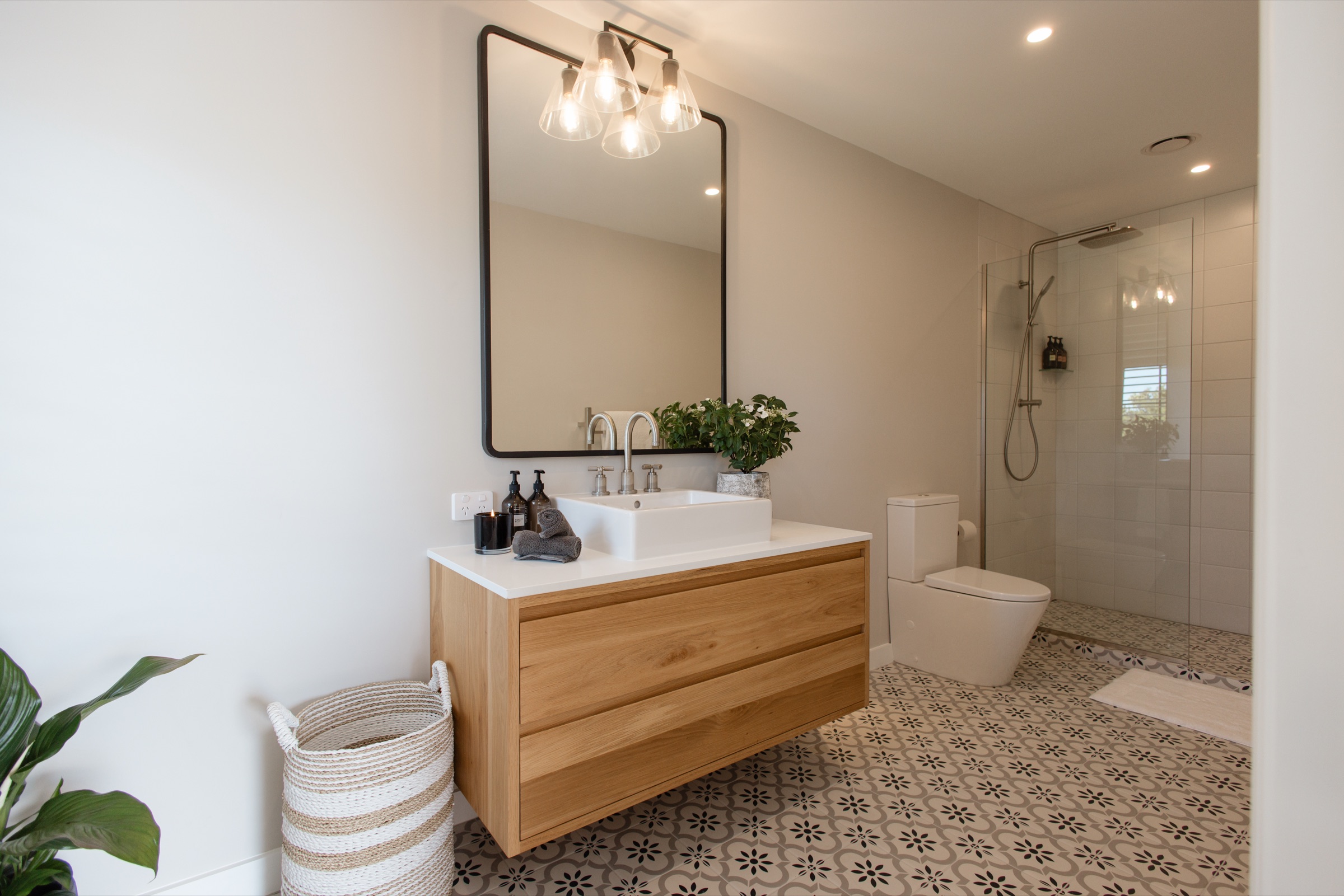 A bathroom in the Farmhouse at Bluestone Ridge: a floating oak vanity with a white basin, a black-framed mirror with pendant lights above, patterned tiled floor, and a glass walk-in shower at the end.