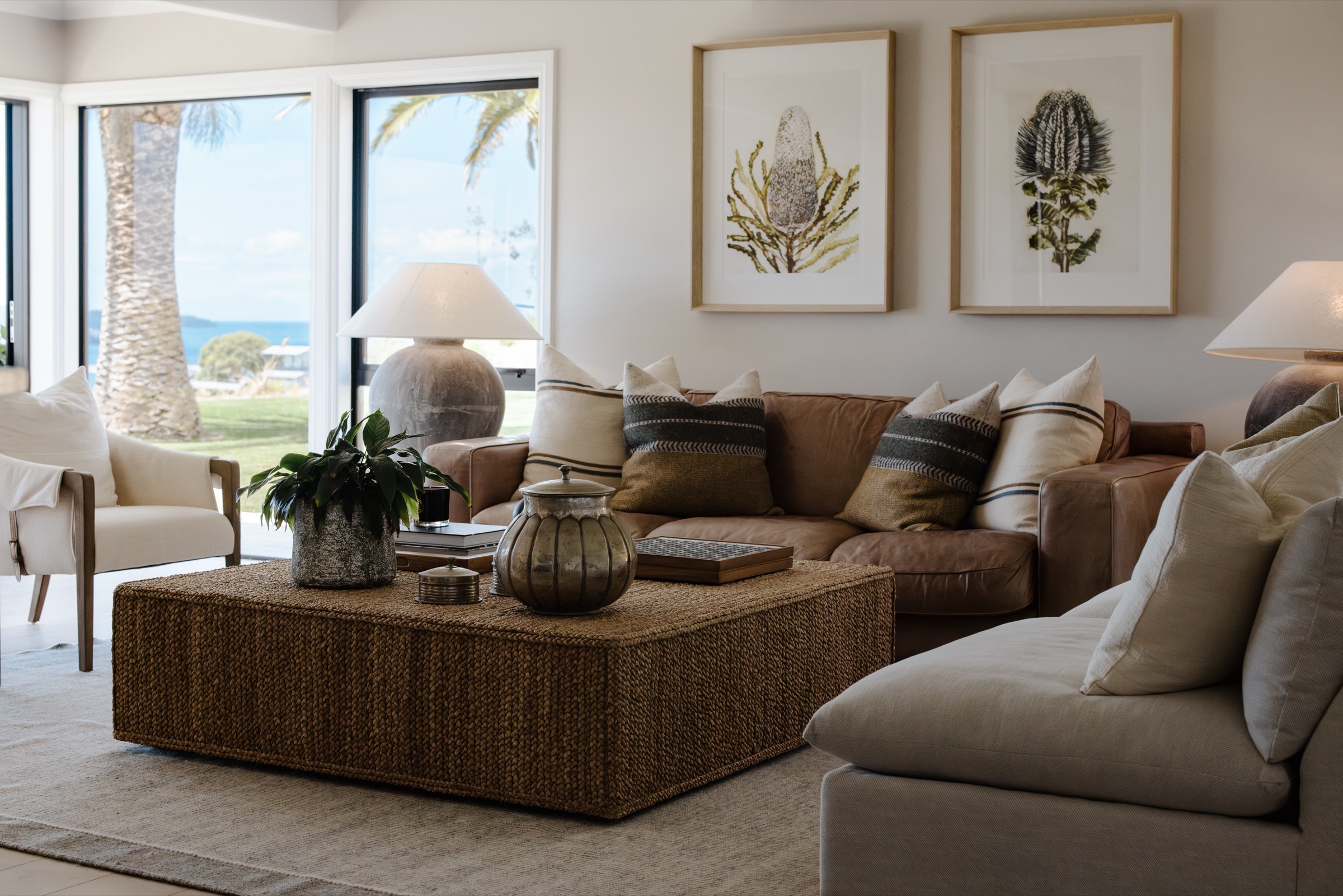 The Farmhouse living room at Bluestone Ridge: tan leather sofa with striped cushions, a woven seagrass coffee table, botanical prints, and palms and the sea through the windows.