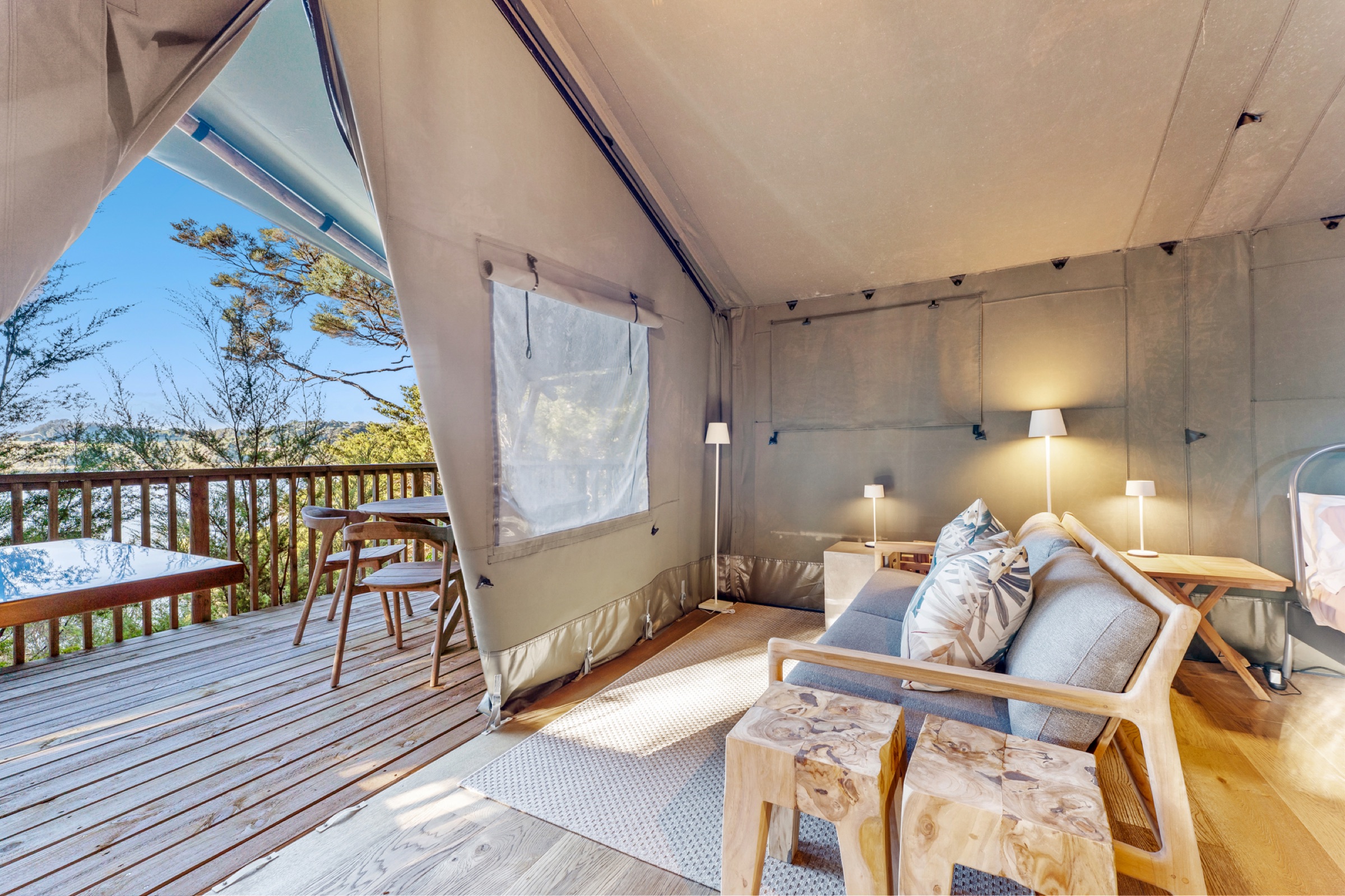 Inside Safari Tent 2 at Bluestone Encampment: a grey sofa with cushions, timber stools, lamps, and the canvas front tied open to the deck, the table, and the estuary through the trees.